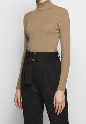 Femme portant un col roulé côtelé beige ajusté et un pantalon noir taille haute avec une ceinture en tissu dotée d'une boucle double anneau dorée.