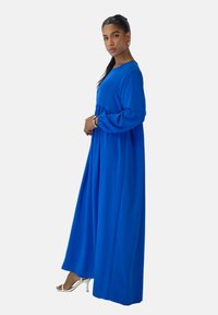 Lange blauwe jurk met een geplooide taille, lange mouwen en een vloeiende silhouet. Draagt witte sandalen met hoge hakken. Gladde stoftextuur.