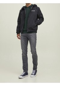 Svart zip-up jacka med huva och logotyp, grön hoodie under, grå jeans och marinblå sneakers med vita sulor. Avslappnad outfit visad.