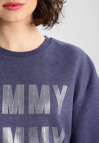 Tmavě modrá mikina s kulatým výstřihem, s lesklým kovov�ým stříbrným textem "MY" v pruhovaném designu. Měkká textilie.
