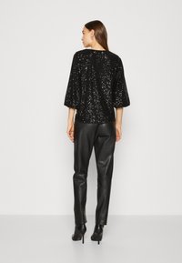 Schwarzes, mit Pailletten besetztes Oberteil mit dreiviertel Ärmeln, kombiniert mit schwarzen Lederhosen und schwarzen Stiefeletten mit Absatz. Glatte Textur; glänzende Oberfläche durch Pailletten.