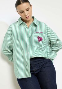 Groen en wit gestreept oversized shirt met knoopsluiting, voorzien van een roze sequined hartpatch en geborduurde tekst op de borstzak.