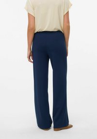 Pantalons larges bleu marine avec une texture lisse, taille haute et coupe ajustée. Mannequin portant un haut beige clair et des chaussures beiges.