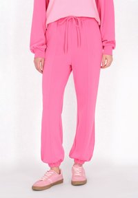 Rózsaszín sportnadrág, húzózsinórral a derékrészen és gumis mandzsettákkal. Simább anyag, oldalsó varrással. Világos rózsaszín edzőcipőkkel párosítva.