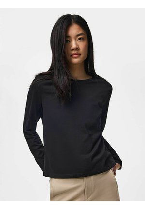 Jeune femme aux longs cheveux noirs portant un haut noir à manches longues et un pantalon beige, debout devant un fond clair uni.