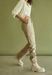 Vrouw in beige cargobroek, witte cropped top en dikke off-white laarzen, wandelend tegen een olijfgroen achtergrond.