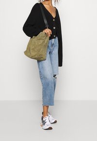 Olivgrön mocka axelväska med mjuk textur, ihopparad med en svart stickad cardigan, vissna ljusblå jeans och vita träningsskor.