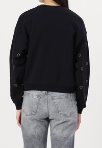Personne portant un sweat-shirt noir avec de petits motifs de cœurs blancs sur les manches et un jean gris délavé, vue de dos.