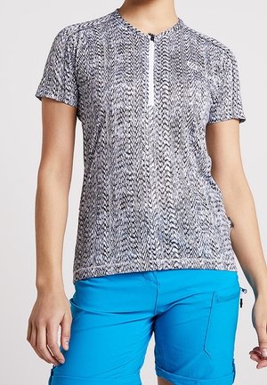 Kvinde iført sort-hvid mønstret kortærmet lynlåstrøje og klareblå korte shorts med lynlåslomme, stående foran en ensfarvet baggrund.