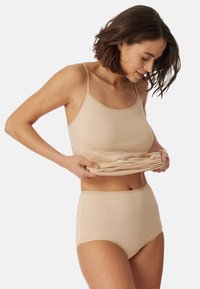 Beige tank top ja korkeat vyötäröiset alushousut, jotka on valmistettu sileästä, joustavasta kankaasta. Tankissa on ohutolkaimet ja pyöreä kaula-aukko.