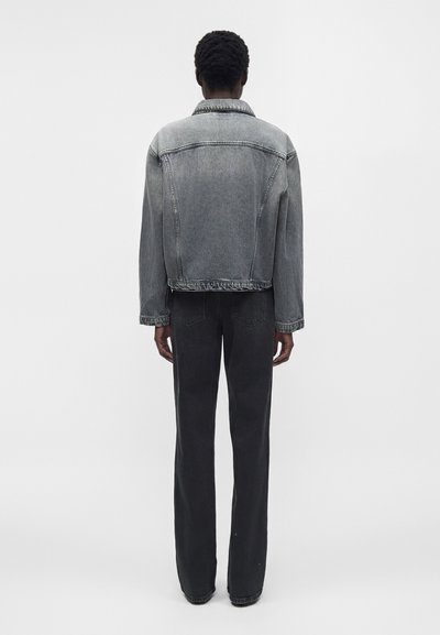 Άτομο που φορά ξεθωριασμένο μαύρο τζιν μπουφάν και μαύρο τζιν με ψηλή μέση, στέκεται με την πλάτη σε απλό λευκό φόντο.