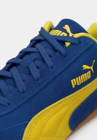 Blauwe suède sneakers met gele accenten. Voorzien van platte veters, een gevoerde kraag en Puma-logo. Textuurpaneel aan de zijkant en rubberen zool.
