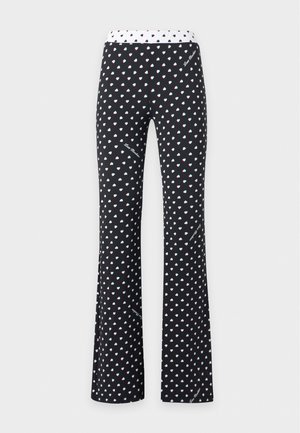 Femme portant un pull rose Love Moschino, un pantalon noir avec des motifs de cœurs blancs et des sandales à talons noires ouvertes, debout avec une main sur la hanche.