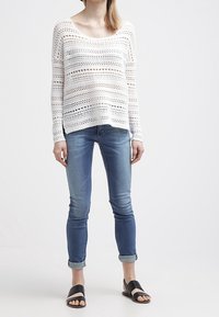 Pull en maille blanche à tissage ouvert, col rond, manches longues, associé à un jean skinny bleu et des sandales noires à enfiler.