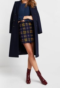 Námořnický dlouhý kabát přes tmavě modrou blůzu, spárovaný s károvanou mini sukni v námořnické modři, žluté a bílé. Burgundské kotníkové boty dotvářejí vzhled.