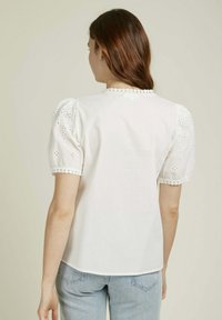 Blouse blanche avec des manches bouffantes mettant en valeur des détails en dentelle et des broderies. Tissu en coton doux avec un col rond et un ourlet festonné.