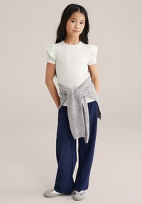 Top bianco a maniche corte con dettagli arricciati, fascia grigia in maglia annodata in vita, pantaloni larghi blu navy e scarpe grigio chiaro.