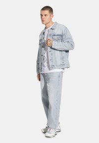 Jasnoniebieska kurtka dżinsowa z podwójnymi kieszeniami na piersiach, w zestawieniu z jasnoniebieskimi dżinsami. Model nosi białą koszulkę i białe buty sportowe.