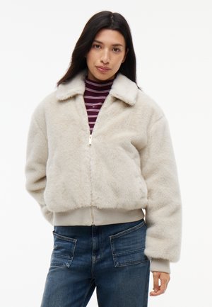 Woman with long dark hair wearing a cream zip-up furry jacket over a striped burgundy turtleneck and blue jeans standing against white background.