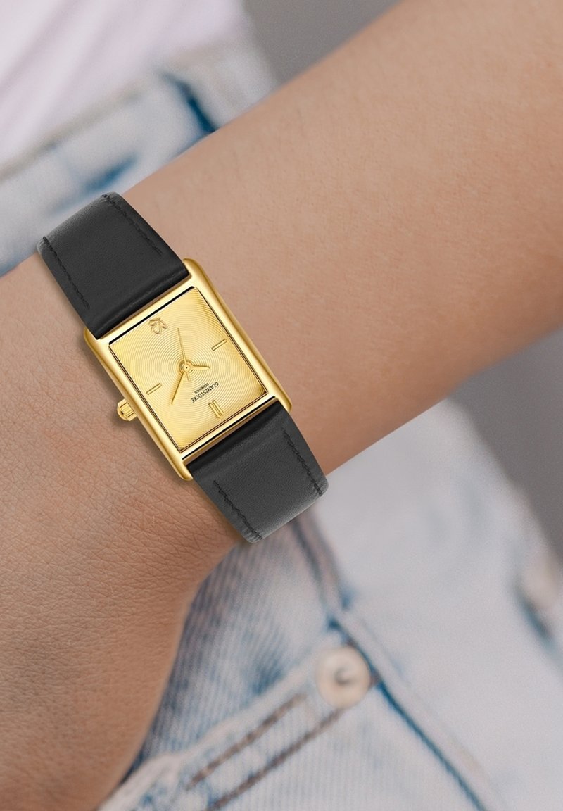 Reloj rectangular dorado con un dial texturizado, correa de cuero negro y marcadores de hora simples en una muñeca contra una tela de mezclilla.