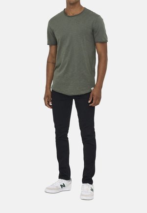 Mann trägt olivgrünes kurzärmeliges T-Shirt, schwarze eng anliegende Hose und weiße Sneaker mit grünem "N"-Logo, steht vor einfachem Hintergrund.