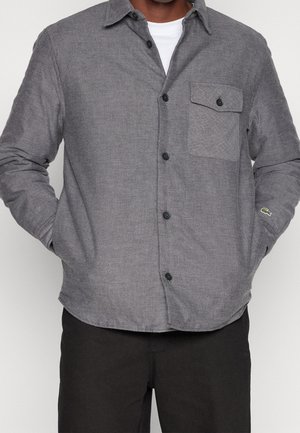 Mand iført en grå skjorte med brystlomme og sorte knapper, hænderne i lommerne, kombineret med sorte bukser.