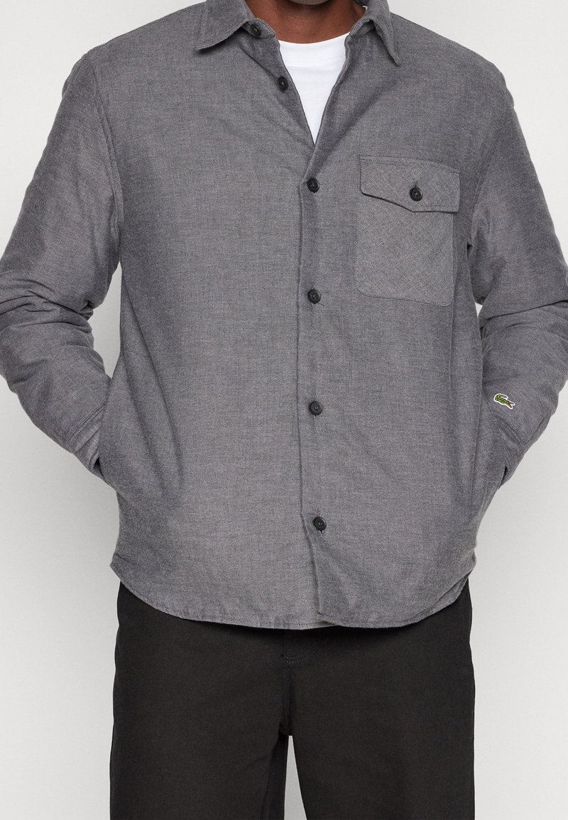 Homme portant une chemise grise boutonnée avec une poche poitrine et des boutons noirs, les mains dans les poches, associée à un pantalon noir.