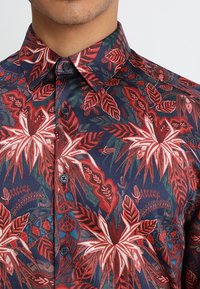 Chemise à manches courtes avec un fond bleu foncé, ornée de grands motifs floraux rouges et blancs, col boutonné et boutons noirs.