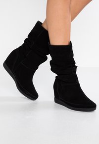 Bottes compensées en daim noir au design décontracté, avec une texture lisse et un bout arrondi et bas. La semelle est plate et subtilement profilée.