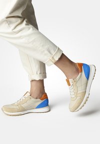 Sneaker beige con pannelli in suede testurizzato, dettagli blu e arancioni, lacci bianchi e suola in gomma. Progettato con un profilo basso e una forma arrotondata.
