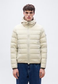 Νέος άνδρας που φοράει μπεζ καπιτονέ μπουφάν με κουκούλα και μπλε τζιν, στέκεται μπροστά σε απλό λευκό φόντο.