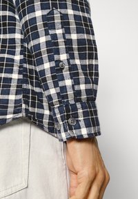 Blue and white checkered button-up shirt with a soft texture, featuring two buttons at the cuff and a pointed collar.