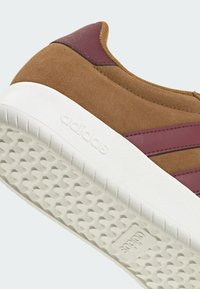 Brauner Wildleder-Sneaker von Adidas mit bordeauxroten Streifen und Fersenakzent, weiße strukturierte Sohle mit eingeprägtem Adidas-Logo.