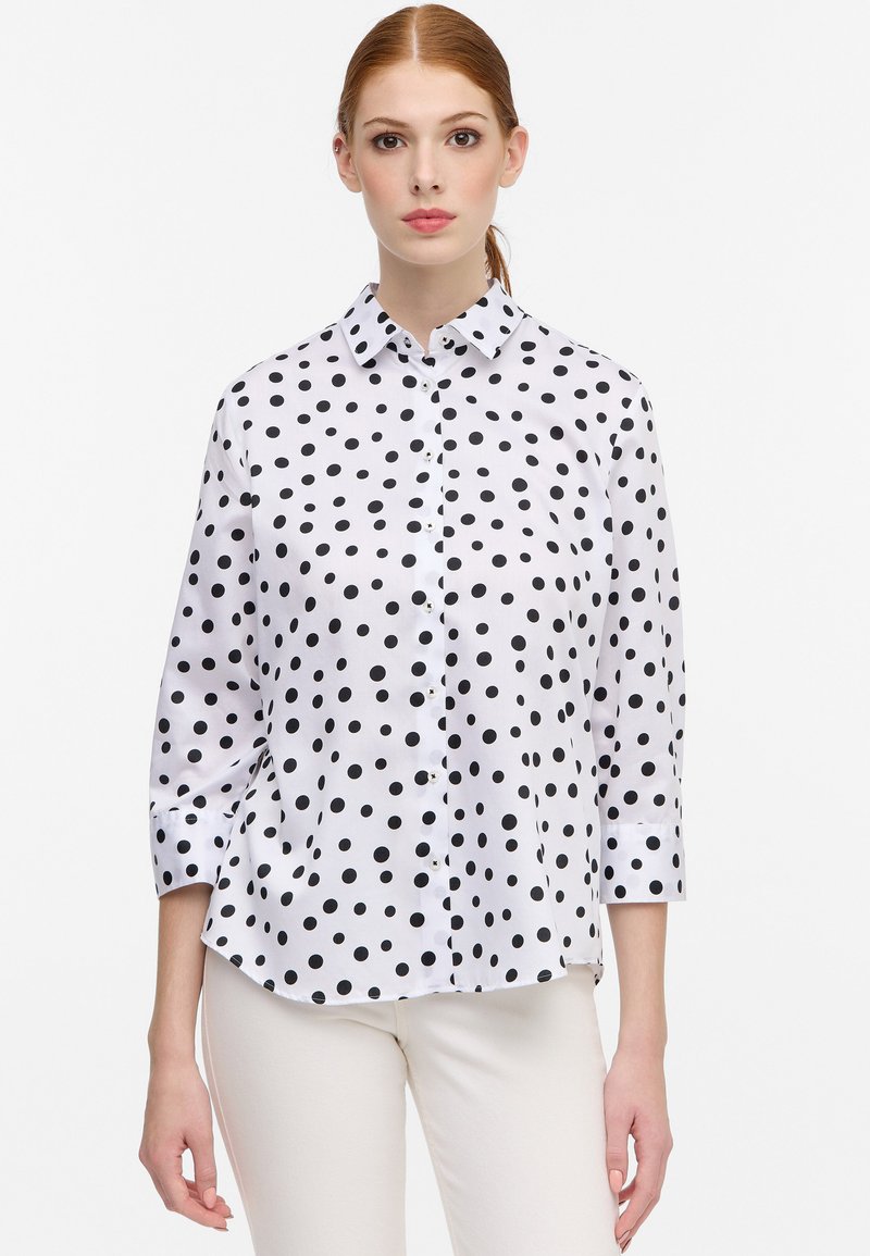 Witte blouse met zwarte stippen, met een kraag en driekwart mouwen. Soepele stof, klassieke pasvorm.