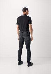 Camisa polo negra con detalles blancos, combinada con jeans oscuros de corte slim. El modelo está de pie, mostrando la vista trasera del atuendo. Zapatos negros completan el look.