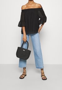 Top nero scollato sulle spalle con maniche a strati, abbinato a jeans blu chiaro a gamba larga. Tiene in mano una piccola borsa a tracolla nera intrecciata. Sandali neri con cinturini.