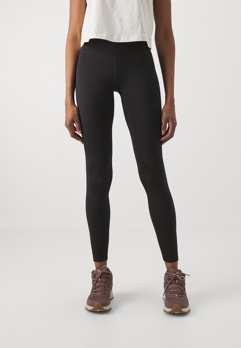 Leggings neri realizzati in tessuto elastico, con vita alta, superficie liscia e design aderente. Indossati con scarpe da trekking marroni.