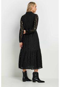 Svart spets maxi-klänning med en tiered fåll, långa ärmar och krage, parat med svarta cowboyboots, som visar ett texturerat blommönster.
