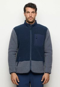 Giacca in pile con colletto alto, blocchi di colore blu navy e grigio, tessuto testurizzato, tasca sul petto con zip e tasche laterali. Fabbricata a macchina.