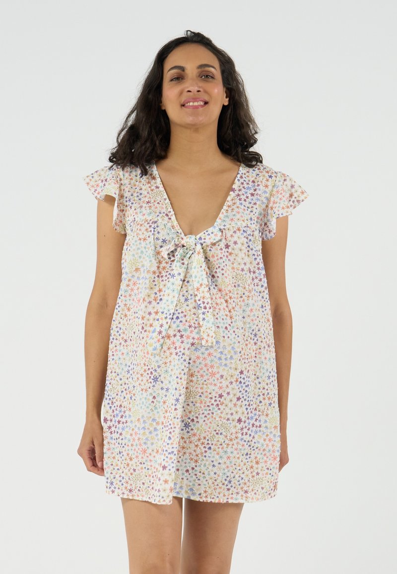 Frau mit dunklem, lockigem Haar trägt ein kurzes, lockeres Kleid mit Blumenmuster, Flatterärmeln und einer vorn gebundenen Schleife, steht vor einem einfarbigen Hintergrund.