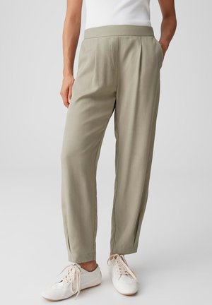 Pantalon plissé taille haute beige porté avec des baskets blanches et un top blanc sans manches, les mains détendues le long du corps.