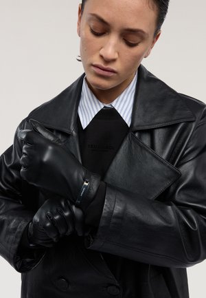 Sorte læderhandsker med en glat tekstur, båret med en sort sweatshirt og en stribet skjorte med krave, sat op mod en neutral baggrund.