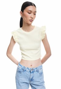 Gul ribbet crop top med flæseærmer. Båret med høj talje lyseblå jeans. Glat tekstur, tætsiddende design og rund halsudskæring.