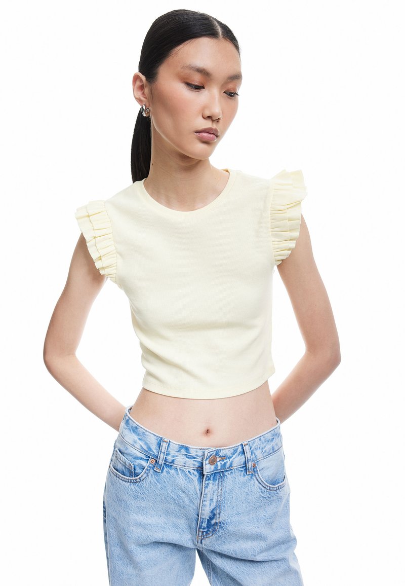Keltainen ribbipinta crop-top, jossa on röyhelöhuput. Yhdistetty korkeavyötäröisten vaaleansinisien farkkujen kanssa. Sileä kangas, napakka malli ja pyöreä pääntie.