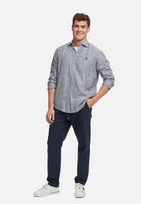 Camicia button-up a righe blu e bianche, abbinata a pantaloni navy scuri e sneakers bianche. Presenta un piccolo logo sul petto.