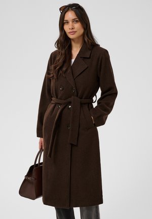 Femme aux longs cheveux foncés porte un manteau en laine marron ceinturé et tient un sac à main marron foncé, avec des lunettes de soleil posées sur sa tête.