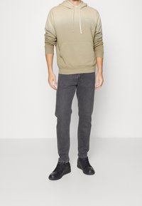 Felpa con gradiente beige, tasca frontale e cappuccio con coulisse, abbinata a jeans grigio scuro e sneaker nere. Design semplice e casual.
