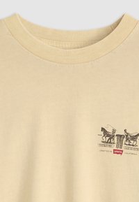 Beige T-shirt met ronde hals en een kleine zwarte grafiek van twee paarden die spijkerbroeken trekken, met het rode Levi's-logo op de borst.