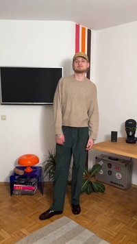 Μπεζ πλεκτό πουλόβερ, άνετη γραμμή, συνδυασμένο με παντελόνια στενής γραμμής σε σκούρο πράσινο. Καφέ δερμάτινα παπούτσια ολοκληρώνουν το ντύσιμο. Μινιμαλιστική διακόσμηση φόντου.