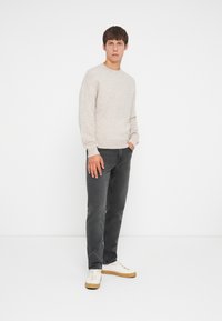 Lys beige striktrøje, mørkegrå jeans og cremefarvede sneakers med brune såler. Modellen står med den ene hånd i lommen mod en hvid baggrund.